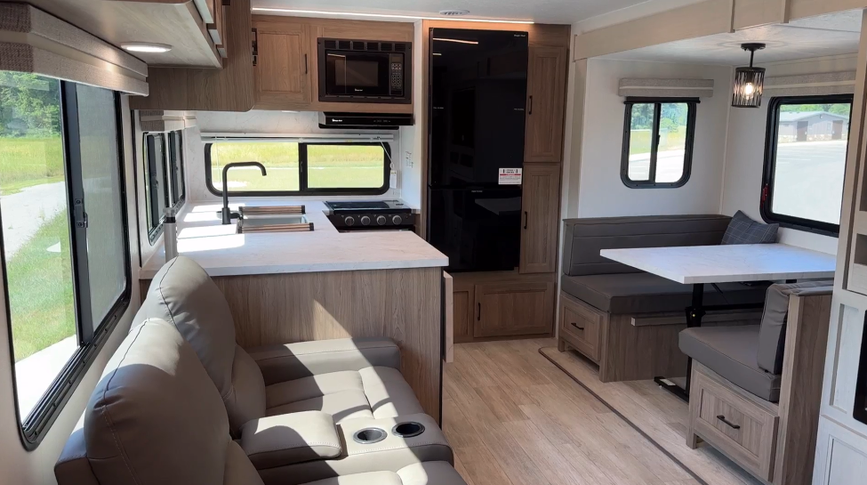 Modern RV interior featuring a spacious layout with a kitchen area, dining booth, and reclining leather seats. The kitchen includes a countertop, sink, and black appliances, while large windows provide natural light.