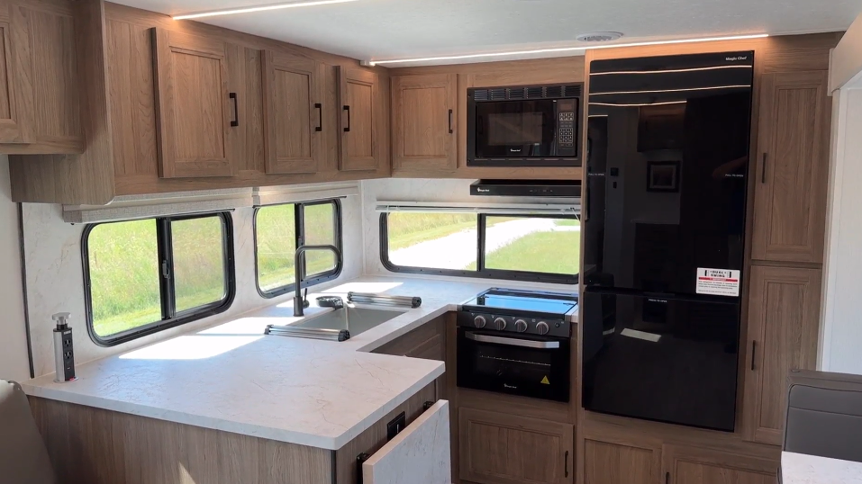 RV kitchen with elegant wooden cabinets, a spacious countertop, a sleek black refrigerator, and built-in appliances, including a stove and microwave. Large windows let in natural light, creating a bright and welcoming cooking space.