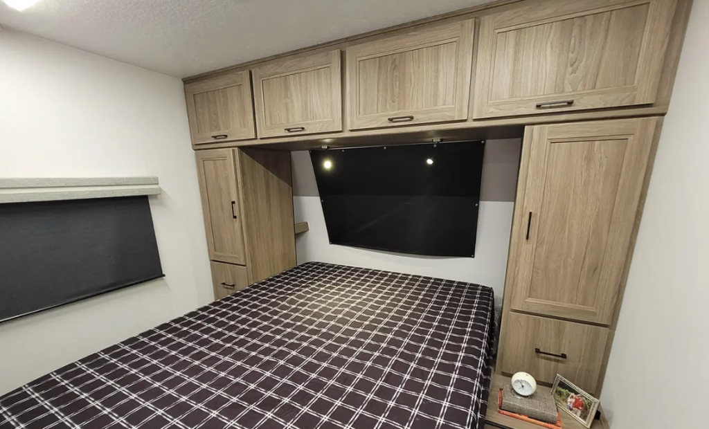 RV bedroom with a cozy bed covered in a checkered pattern and surrounded by overhead and side wooden cabinets for storage. A window with a blackout shade provides privacy, and the room's minimalist design creates a comfortable and functional sleeping area.