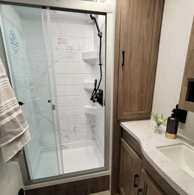 RV bathroom with a modern glass-enclosed shower featuring marble-patterned wall tiles and built-in shelves. The vanity area includes a sleek countertop, under-sink storage, and a wooden cabinet, creating a clean and organized space.
