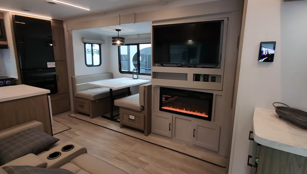 Spacious RV living area featuring a cozy dining booth, a flat-screen TV mounted above a glowing electric fireplace, and a comfortable seating arrangement with plush recliners. The design emphasizes a modern aesthetic with ample natural light and functional storage.