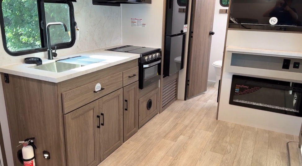 RV kitchen and living space featuring a modern design with a wooden cabinet kitchenette, a stainless steel sink, and a three-burner stove with an oven. Adjacent to the kitchen, a refrigerator and microwave offer additional functionality. The space also includes a flat-screen TV above an electric fireplace, with a door leading to a bathroom in the background. Large windows and light-colored flooring enhance the open, airy feel.