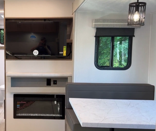 RV interior showcasing an entertainment area with a mounted flat-screen TV above a built-in electric fireplace. To the right, a dining nook features a marble-style table and cushioned seating beneath a modern pendant light. A nearby window brings in natural light, enhancing the cozy yet contemporary design.