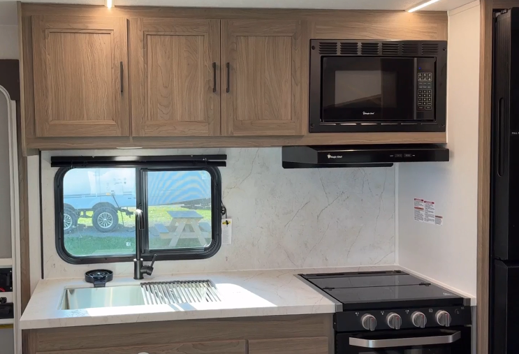 RV kitchen featuring a compact and efficient design with wooden overhead cabinets, a microwave, and a three-burner stovetop with an integrated oven. A marble-style countertop includes a stainless steel sink positioned under a window, providing natural light and a view of the outdoors. The backsplash has a clean, modern look, emphasizing practicality and style in a small space.