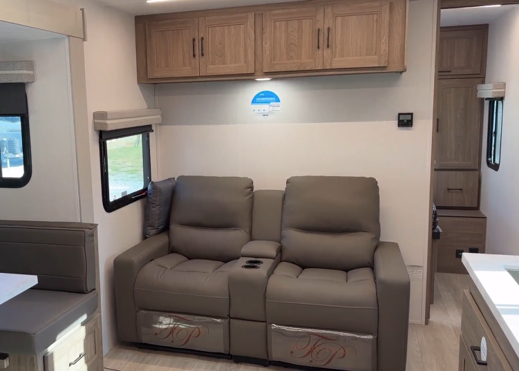 Interior view of an RV living space featuring a pair of reclining seats with cup holders in a modern, neutral color palette. Above the seats are wooden storage cabinets, and natural light enters through the side windows. The design highlights comfort and functionality for on-the-go living.