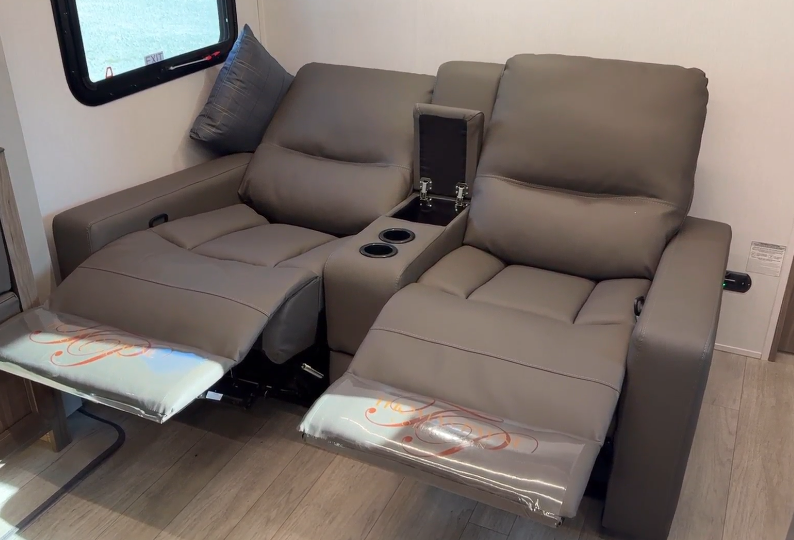 Interior of an RV featuring two fully reclined theater-style seats with leg rests extended. The plush brown leather upholstery includes cup holders and a center console with a hidden storage compartment. A nearby window provides natural light, emphasizing the cozy and functional design.