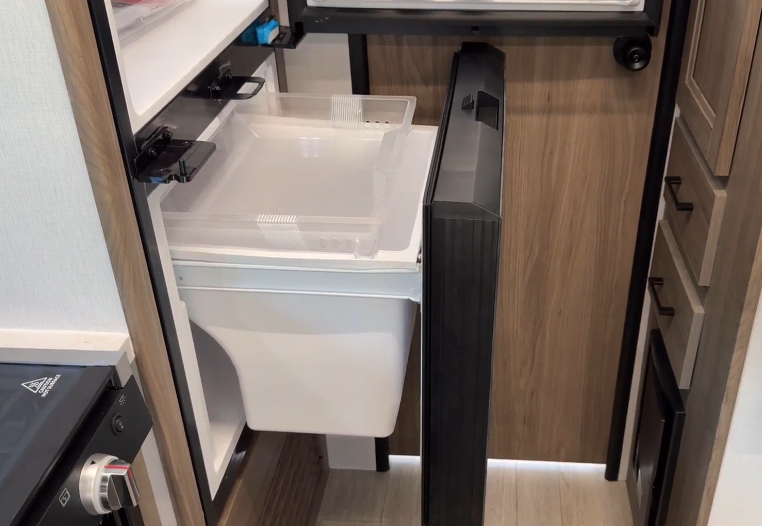 Open RV refrigerator showcasing its spacious interior with organized compartments, including a clear drawer for storage and a large bottom bin for bulky items. The sleek black door contrasts with the warm wood paneling, emphasizing a functional and stylish design in the compact kitchen area.