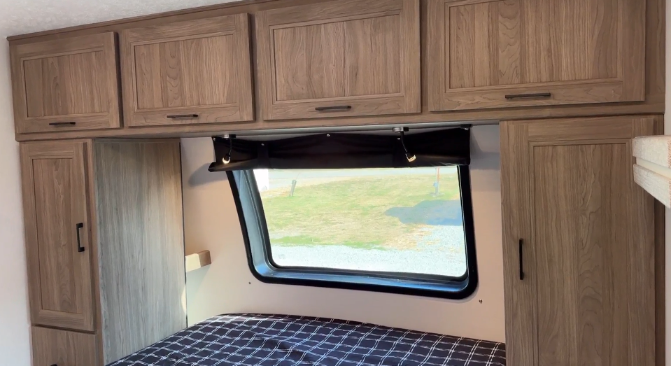  RV bedroom featuring a cozy design with a large window framed by wooden overhead cabinets and side closets for ample storage. Below the window, a neatly made bed with a plaid-patterned bedspread adds a touch of comfort. Natural light from the window brightens the space, enhancing its warm and practical layout.