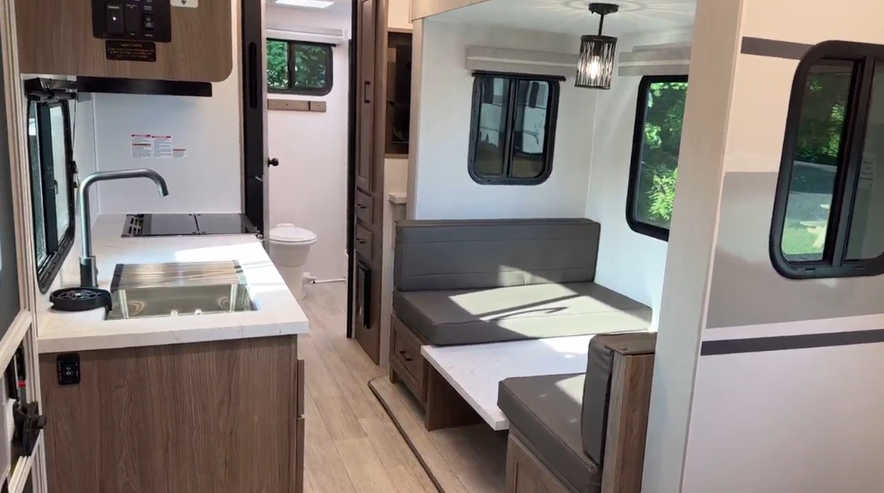  Open-concept RV interior showcasing a modern kitchen with a stainless steel sink and wooden cabinetry on the left. The dining area features a convertible dinette with cushioned benches and a marble-style tabletop under a stylish pendant light. Large windows allow natural light to fill the space, enhancing the bright and airy design. A hallway leads to a bathroom visible in the background, completing the practical and cozy layout.