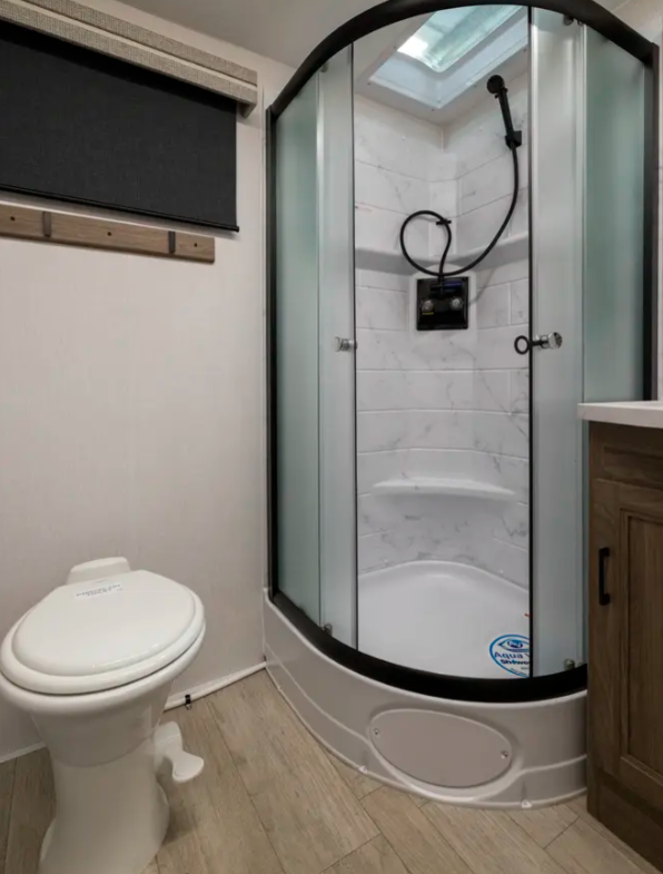 RV bathroom featuring a compact design with a curved glass shower enclosure, complete with a marble-style interior and built-in seating. A skylight above the shower provides natural light, while a modern white toilet is positioned nearby. A wooden vanity with a countertop adds storage and functionality, complementing the sleek and practical layout.