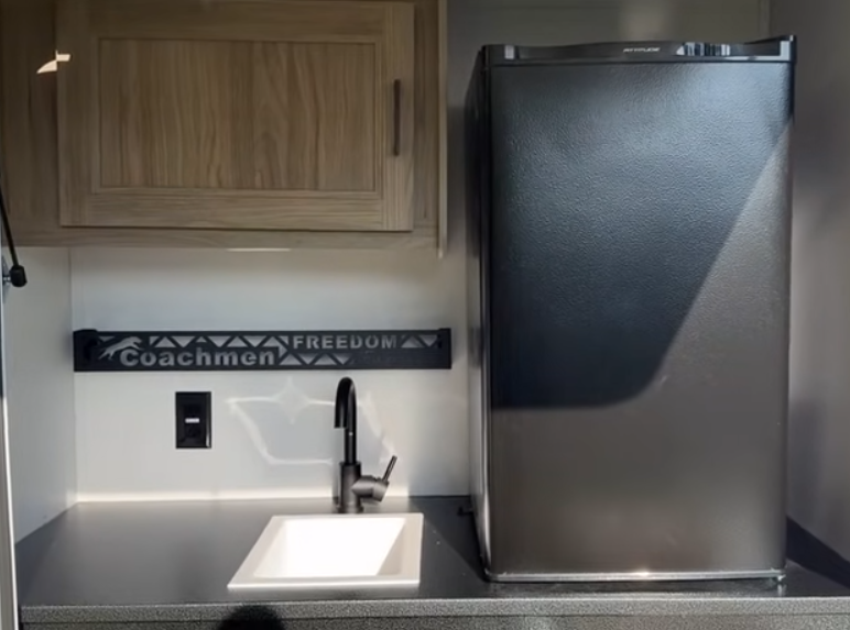 A well-designed outdoor camp kitchen featured in the Freedom Express 252RBS. Key elements include a compact refrigerator, a real sink with a faucet and drain, and upper storage cabinetry. 