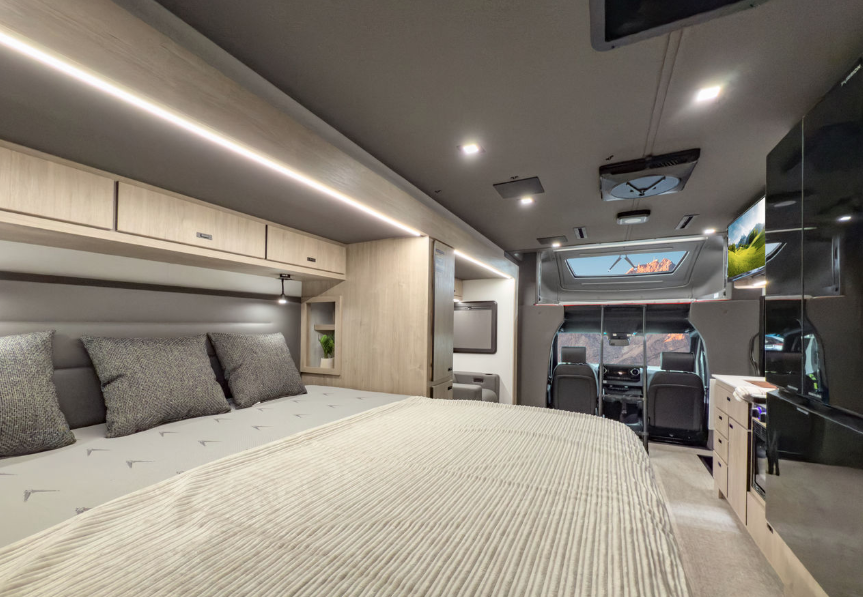 RV interior showcases a luxurious sleeping area with a spacious bed surrounded by sleek light wood cabinetry. The overhead storage and bedside compartments offer practical organization solutions, complemented by ambient LED lighting that enhances the modern aesthetic. The layout flows into the cab area, where a skylight and expansive windshield provide natural light and scenic views. Thoughtful design elements, such as a built-in shelf and soft gray tones, create a cozy yet sophisticated environment, perfect for restful nights on the road.