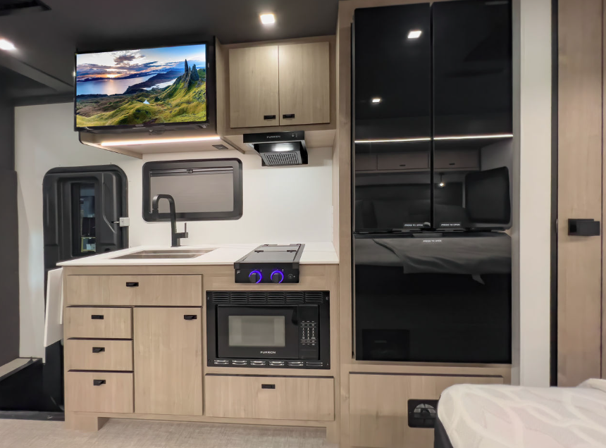 This RV kitchen area exudes modern functionality with its sleek design. The light wood cabinetry pairs seamlessly with a solid white countertop, creating a clean and inviting space. The black appliances, including a microwave, stovetop, and double-door refrigerator, add a bold contrast. A mounted flat-screen TV enhances entertainment options, while the minimalist faucet and backsplash elevate the aesthetic. Ample storage is provided by multiple cabinets and drawers, ensuring practicality during travel.
