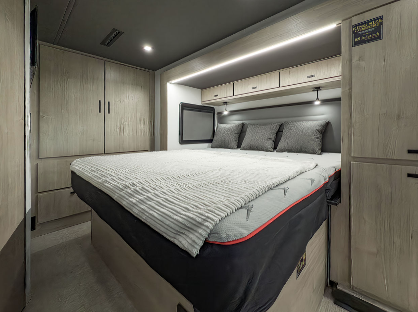 This bedroom area within the RV boasts a king-sized bed, perfectly complemented by neutral tones and clean lines for a contemporary feel. The surrounding cabinetry provides ample storage for extended travels, while the overhead reading lights add a touch of practicality and ambiance. The sleek wood finishes and integrated drawers offer a cohesive and modern aesthetic, ensuring both functionality and style. This thoughtfully designed space prioritizes comfort and convenience, creating a perfect retreat after a day of adventure.