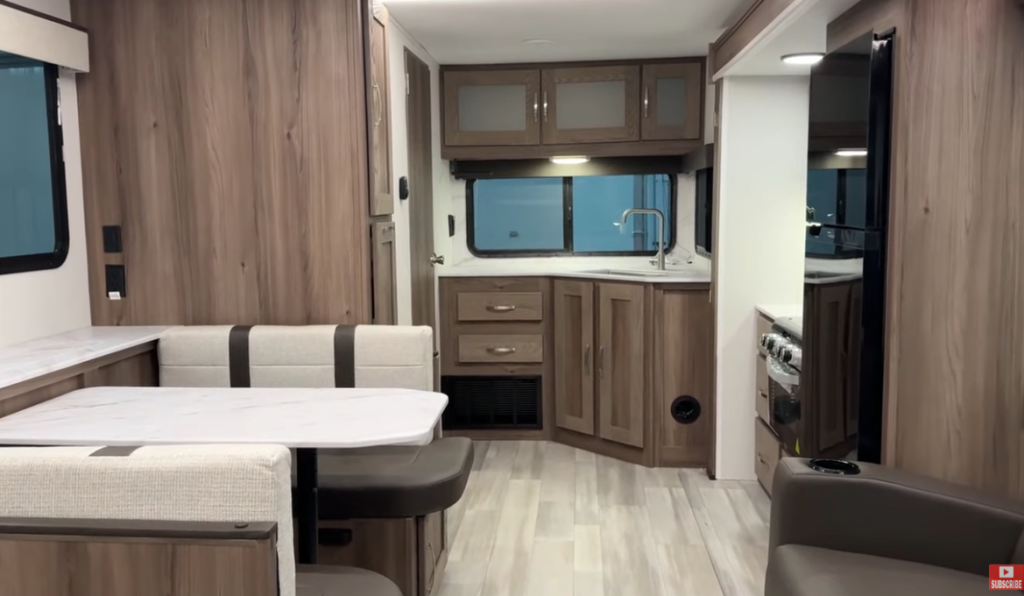 Spacious interior of a GO Explore travel trailer featuring a modern dining area with a bench-style dinette, a corner kitchen with wood cabinetry, and a sleek stainless steel faucet. The design highlights ample storage, natural wood finishes, and vinyl plank flooring, creating a warm and functional living space for travel and relaxation.