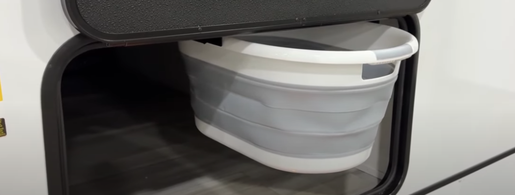 Storage compartment in a GO Explore travel trailer featuring a collapsible gray and white laundry basket, demonstrating efficient use of space and thoughtful organization. This practical addition is ideal for keeping travel essentials neatly stowed away.