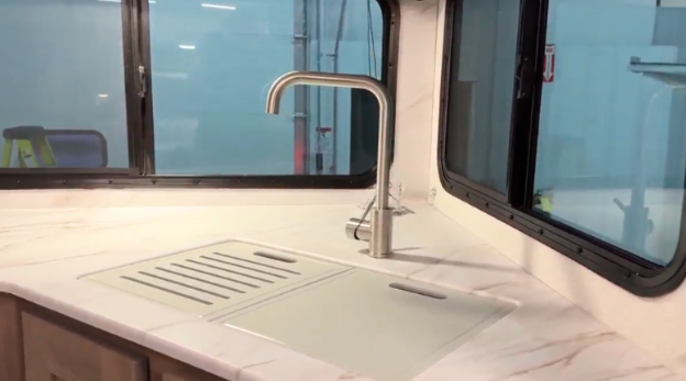 Modern kitchen sink in a GO Explore travel trailer featuring a sleek stainless steel faucet and an integrated cutting board cover. The marble-look countertop and large surrounding windows provide a bright and functional workspace, combining style and practicality for travel living.