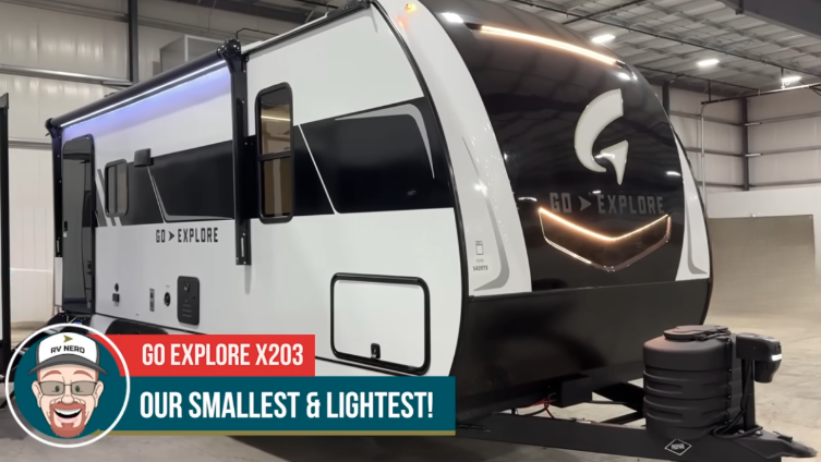 Exterior of the GO Explore X203 travel trailer, showcasing its sleek, modern design with a black and white finish. Highlighted as the smallest and lightest model, it features a compact build, aerodynamic front cap, and dual propane tanks. The trailer is displayed indoors, emphasizing its innovative and lightweight construction.