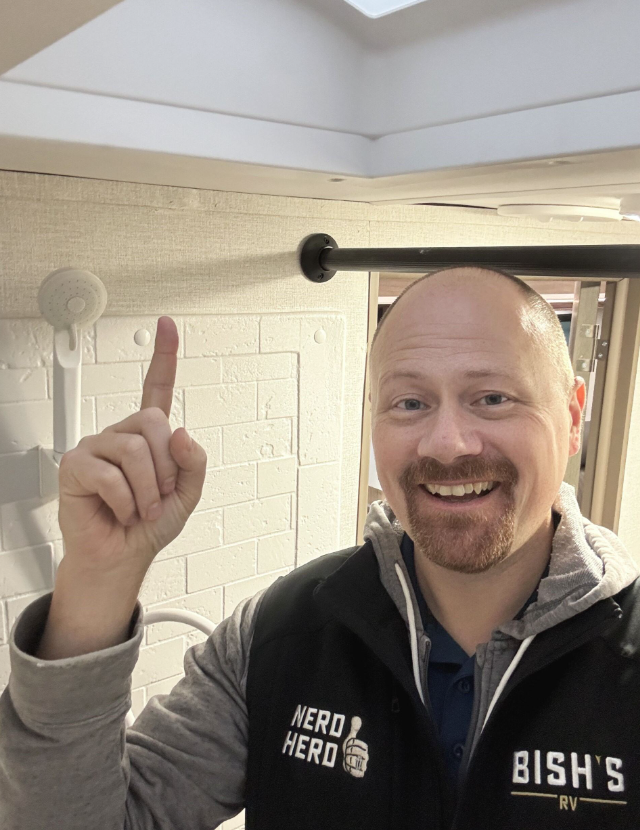 ALT text: A close-up photo taken inside what appears to be the RV bathroom showing a ceiling feature, likely the skylight above the shower mentioned in previous images. The image shows a white ceiling with what looks like a vent or skylight opening. In the foreground is a man with a bald head, reddish-brown beard, and a friendly smile pointing up toward the ceiling feature with his index finger. He's wearing a gray and black jacket with "NERD HERD" and "BISH'S RV" text visible on it. This appears to be the same "RV Nerd" personality seen in the informational overlay from a previous image, likely demonstrating or explaining features of the RV 