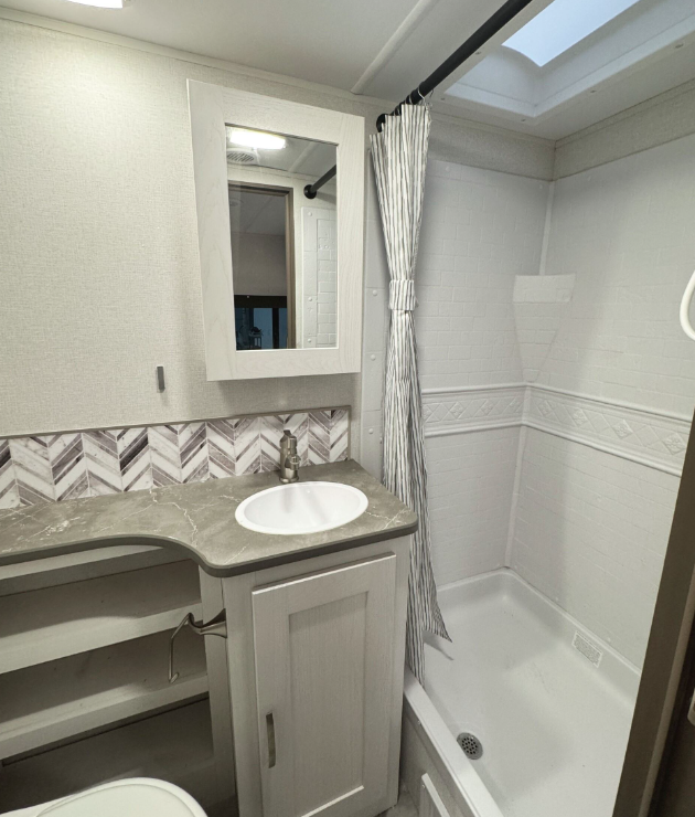  A more complete view of the RV bathroom showing both the shower and vanity area. The bathroom features a white shower stall with white subway-style tile walls and a decorative border, with a striped shower curtain on a black rod. The shower has a skylight above it allowing natural light in. The vanity includes a white cabinet with a gray marbled countertop, white oval sink, and a simple metal faucet. Above the sink is a white-framed mirror with built-in lighting. The vanity backsplash has a chevron pattern in gray and white tones that coordinates with the countertop. The bathroom walls are white with textured surfaces, creating a clean, bright appearance. A small portion of what appears to be the toilet is visible at the bottom left of the frame. The compact bathroom efficiently combines all necessary facilities within the limited space of the RV while maintaining the modern aesthetic seen throughout the vehicle.RetryClaude can make mistakes. Please double-check responses.