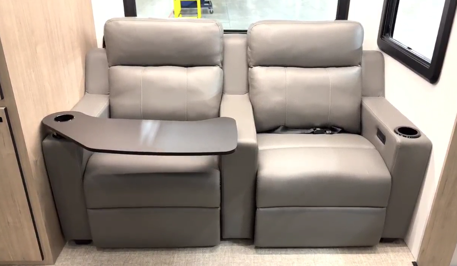 Interior view of a luxury RV or motorhome featuring theater-style seating. The image shows two plush light gray leather recliners positioned side by side in what appears to be a dedicated entertainment area. The left seat has a fold-out table or tray table extended from the armrest, ideal for dining or using devices. Both seats have padded headrests and appear to be recliners. The armrests appear to include cup holders. The seating is installed against light wood-toned wall paneling, matching the modern aesthetic seen in previous images of the high-end motorhome.