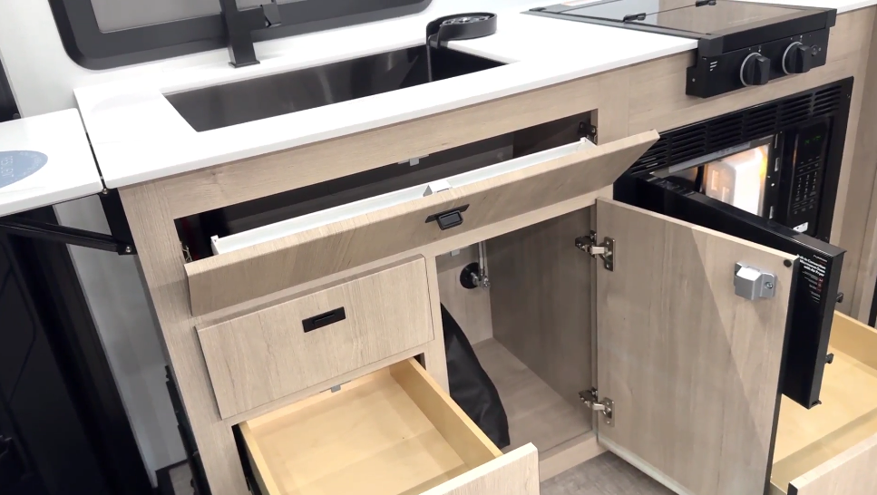 Close-up view of the luxury motorhome's kitchen storage features with multiple compartments open. The kitchen has a white solid surface countertop with a black rectangular undermount sink. The cabinetry is light wood-toned and includes several clever storage solutions: an upper false drawer front below the countertop is hinged and open, revealing a hidden storage area; a lower drawer is pulled out showing a shallow wooden storage compartment; and a cabinet door on the right is open, displaying what appears to be a trash container or storage area inside. On the countertop at right are black control knobs for the cooktop. A black microwave oven with its door open is visible on the right side. 