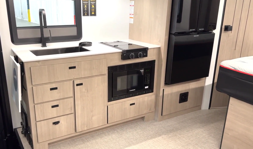 View of the kitchen area in the luxury Lineage motorhome. The kitchen features a sleek, modern design with light wood-tone cabinetry and black hardware throughout. The countertop is white with a black undermount sink and matching black faucet. The cooking area includes a two-burner induction or electric cooktop with black control knobs. Below the counter is a black built-in microwave or convection oven. The cabinetry includes multiple drawers and cabinets with black rectangular pull handles. On the right side of the image is a black refrigerator or appliance tower. A window with a black frame is positioned above the sink area, and what appears to be part of a seating area is visible on the far right edge of the frame. The floor is light gray, continuing the neutral color scheme.