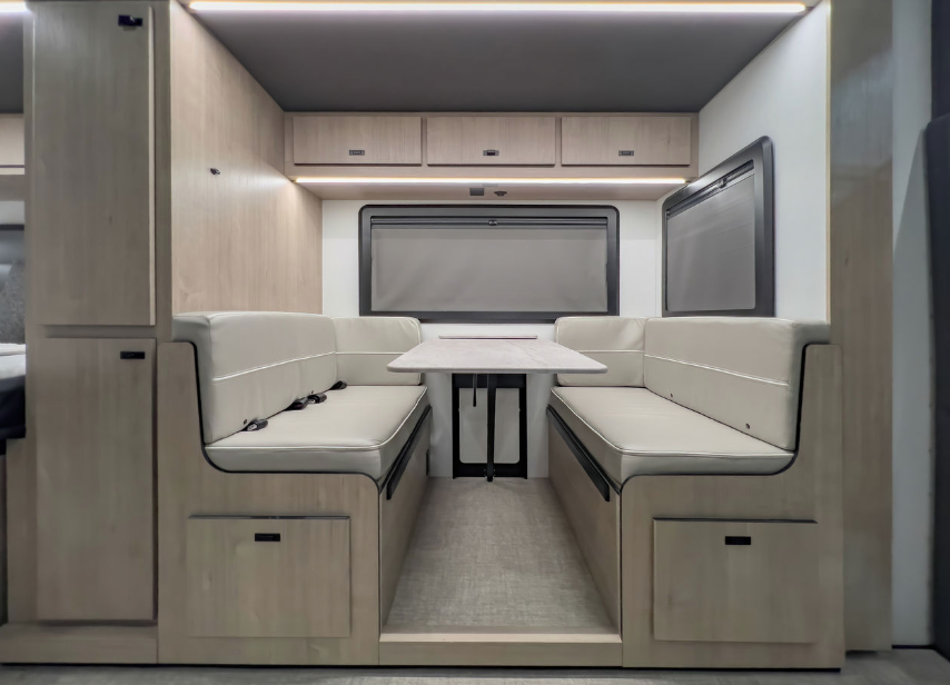  Interior view of a modern RV or motorhome dining area featuring a minimalist, contemporary design. The space has a U-shaped dining booth with white cushioned seating on both sides, accented with black piping or trim. Between the seating is a small white table with a black base. Light wood-toned cabinetry surrounds the dining area, with overhead cabinets at the top and storage compartments beneath the seating, each with black push-to-open handles. The back wall appears to have large windows with privacy shades or covers drawn. The floor has light gray carpeting or flooring. The overall design features a clean, modern aesthetic with a neutral color palette of whites, light woods, and black accents. 