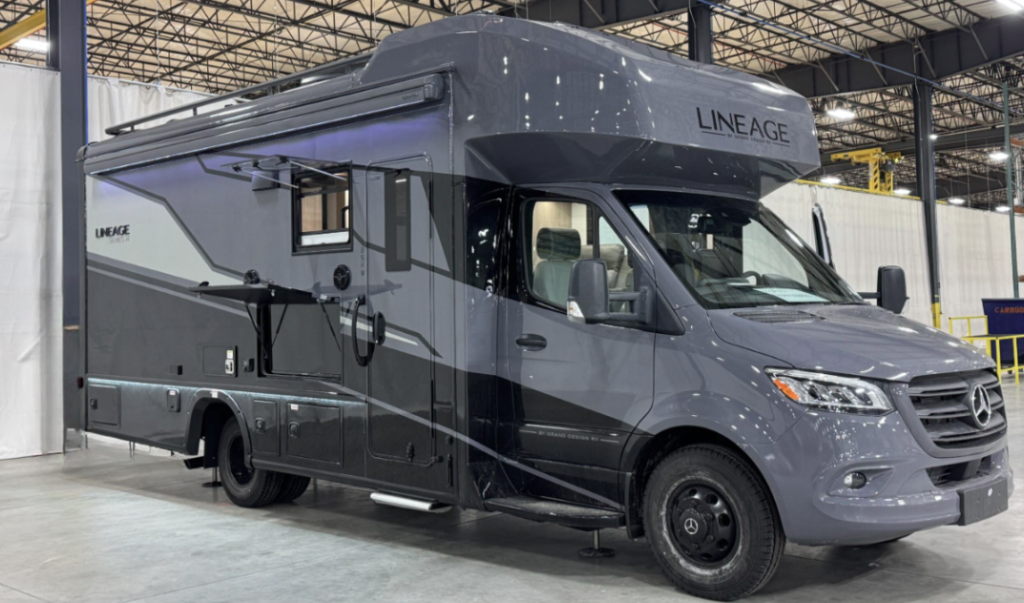 Exterior view of a Class C motorhome or RV branded "LINEAGE" displayed in what appears to be a manufacturing facility or exhibition hall. The motorhome is built on a Mercedes-Benz Sprinter chassis, identifiable by the Mercedes logo on the front grille. It features a sleek, modern design with a predominantly gray exterior with subtle graphic accents. The vehicle has a cab-over design with the living quarters extending over the driving cab, typical of Class C motorhomes. Windows are visible along the side, and there appears to be an awning or slide-out section. The motorhome is parked on a concrete floor inside an industrial space with exposed ceiling trusses and partial wall dividers. This represents a different type of RV from the travel trailers shown in previous images - a self-contained motorhome with integrated driving cab rather than a towable trailer.