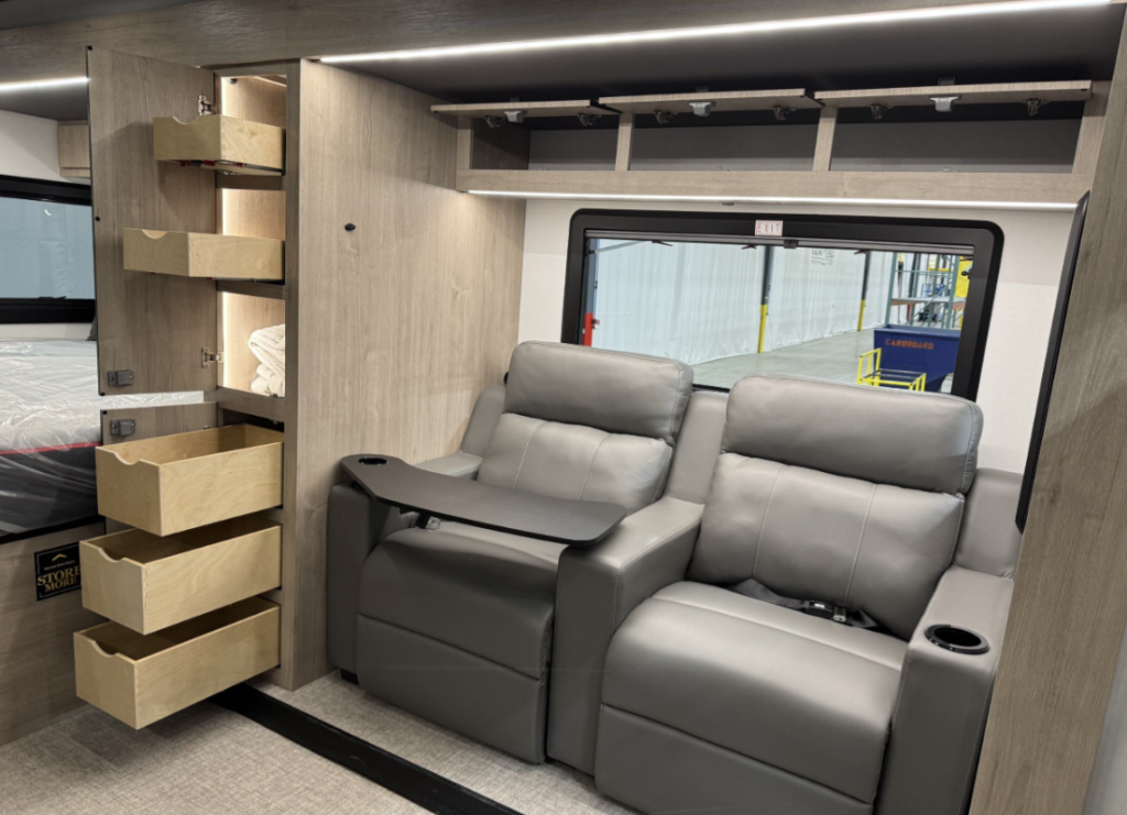 Interior view of the luxury motorhome showing the theater seating area with additional storage features. The same gray leather theater-style recliners are shown, with the left seat having its tray table extended. Behind the seating is a large window with a black frame showing the manufacturing facility outside. On the left side of the image is an impressive storage solution with multiple wooden drawers pulled out at different levels, demonstrating the RV's efficient use of space. The drawers appear to be made of light natural wood, contrasting with the light gray wood-tone exterior cabinetry. The top drawer contains what looks like folded linens or towels. The ceiling features integrated LED lighting strips.