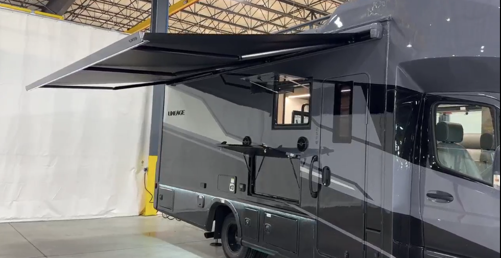 ALT text: Exterior view of the Lineage luxury motorhome from the side, focusing on an extended feature. The image shows the gray motorhome with its large black awning extended from the side, providing an outdoor covered area. The motorhome has the "LINEAGE" branding visible on the exterior. Below the awning, there appears to be an exterior compartment or door open, possibly providing access to outdoor entertainment features or storage. Multiple exterior compartment doors can be seen along the lower portion of the vehicle. The front cab area is partially visible on the right side, showing the Mercedes-Benz Sprinter chassis.