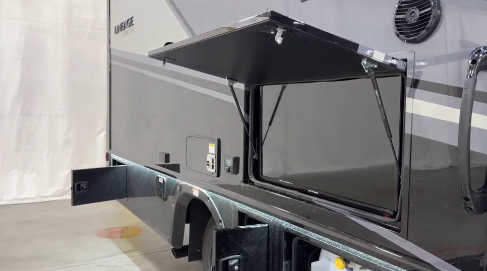 Close-up exterior view of a luxury feature on the Lineage motorhome - an outdoor entertainment system. The image shows a section of the gray exterior of the motorhome with the "LINEAGE" branding visible. A large compartment door is hinged at the top and opened upward, revealing what appears to be a flat-screen TV or entertainment display mounted inside a weatherproof enclosure. Around the TV are various utility connections and control panels. This outdoor entertainment system allows for viewing media while enjoying the outdoors under the awning seen in the previous image. 

