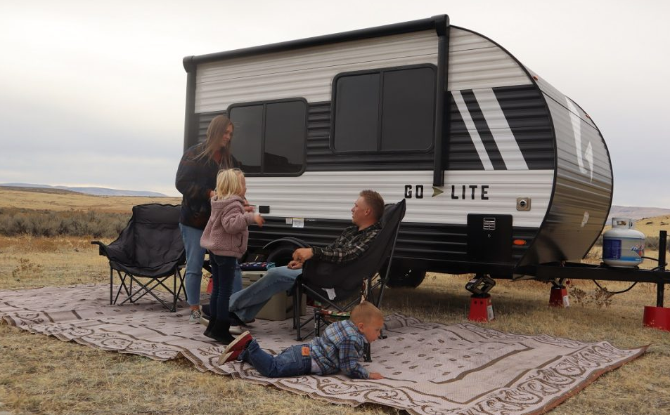 A family camping scene in an open grassland with a "GO LITE" travel trailer parked in the background. The black and white trailer is stabilized on red leveling.