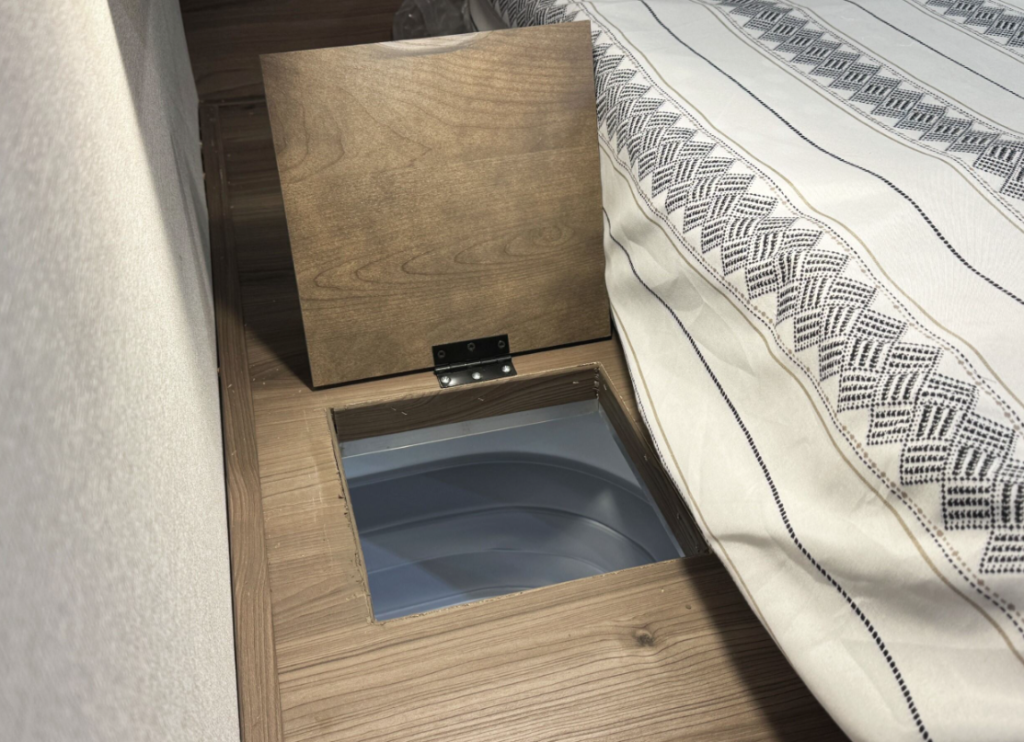 Close-up of an RV bedroom showcasing a hidden storage compartment beneath a wooden hinged panel, located next to a bed with striped geometric bedding. The compartment provides additional space for practical storage solutions within the compact design.