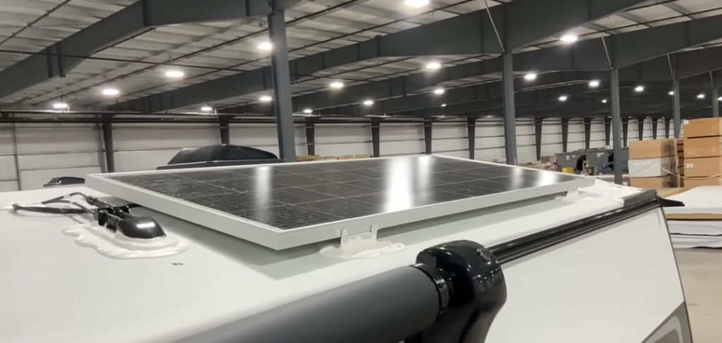  Close-up view of a solar panel mounted on the roof of a travel trailer inside a large warehouse. The panel is securely fastened with brackets and sealed wiring connections, ensuring efficient energy collection. A black awning housing is partially visible in the foreground. The background includes stacks of boxes and other materials, indicating an RV manufacturing or service facility.