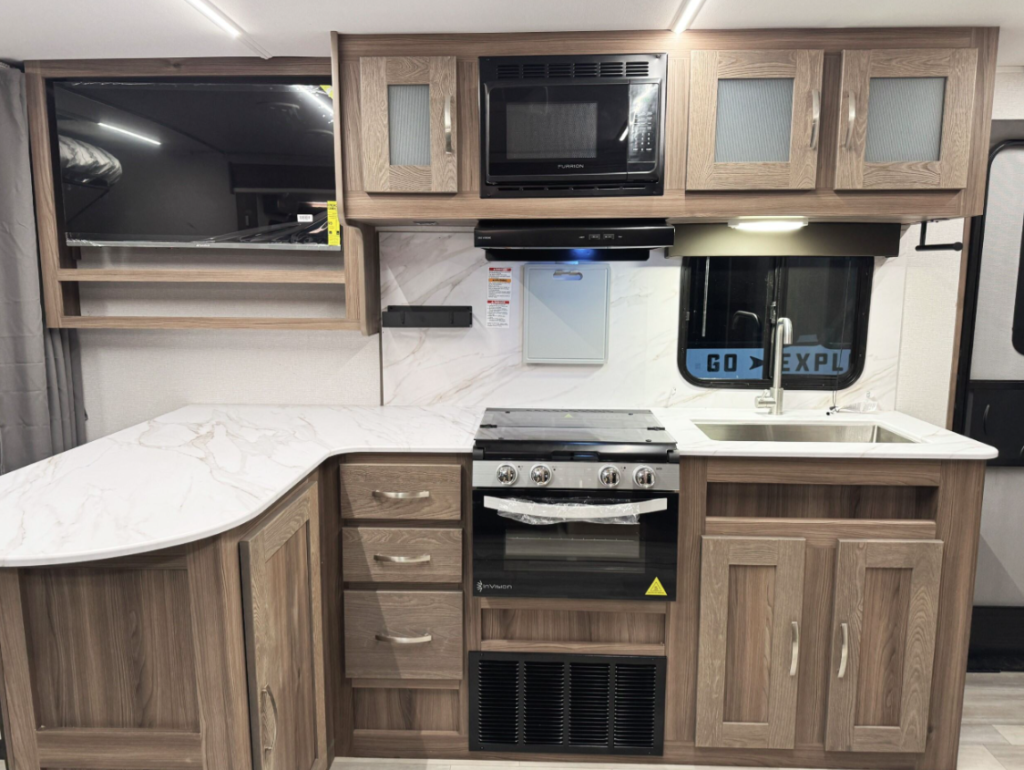RV kitchen interior featuring a U-shaped marble-like countertop, a built-in oven with a stovetop, and wooden cabinetry with silver handles. The upper section includes a microwave and frosted glass cabinet doors, with a mounted flat-screen TV on the left. A stainless steel sink is situated below a window with the words 'GO EXPLORE' visible outside, adding to the modern and functional design.