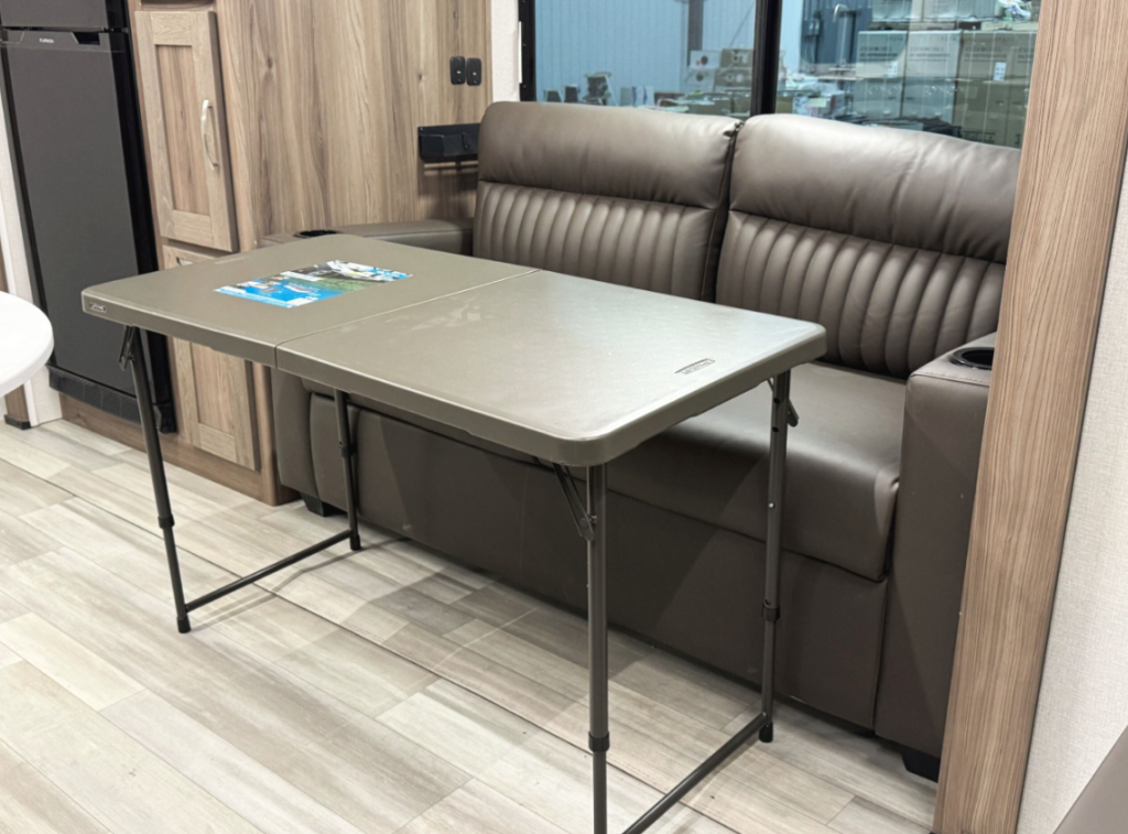 RV interior setup featuring a brown leather loveseat with cup holders, positioned against a large window. In front of the loveseat is a lightweight foldable table with a beige surface, displaying a brochure. The surrounding area includes wooden cabinetry and flooring, adding to the compact and functional design.