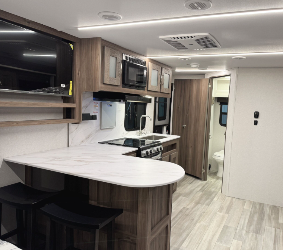 Spacious RV interior featuring a kitchen area with a marble-like curved countertop and bar stools tucked underneath. The kitchen includes a stovetop, oven, microwave, and a stainless steel sink set against wooden cabinetry with frosted glass accents. A flat-screen TV is mounted on the left, and a hallway leads to a bathroom with a visible toilet and additional living space. The light-colored flooring and recessed lighting enhance the modern and open design.