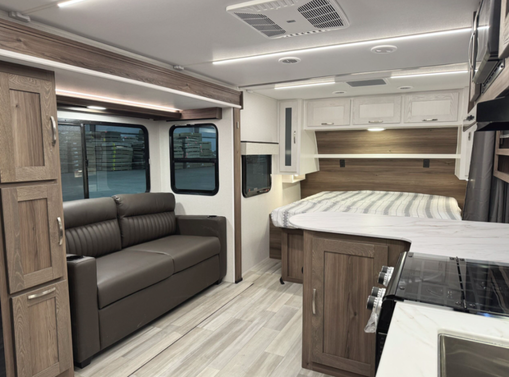 Spacious RV interior featuring a living area with a brown leather loveseat positioned next to large windows. Adjacent is a sleeping area with a bed, striped bedding, wooden paneling, and white overhead storage cabinets. The kitchen area includes a marble-like countertop, stovetop, and sink, with light-colored flooring and recessed lighting throughout for a modern and cozy atmosphere.