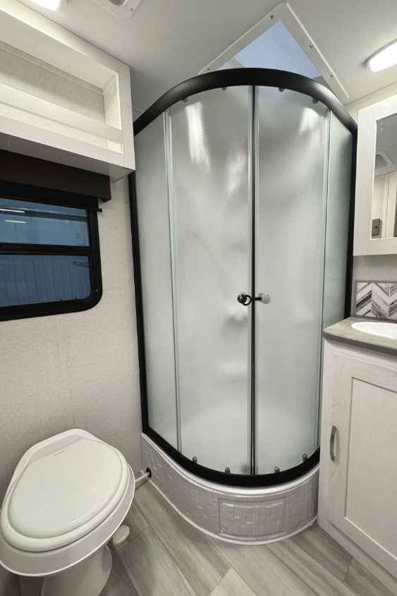 RV bathroom featuring a curved glass shower enclosure with frosted panels, a white toilet, and a vanity with a sink and light-colored cabinetry. The bathroom also includes a window with a pull-down shade, a skylight above the shower, and light woodgrain flooring, creating a bright and functional space.
