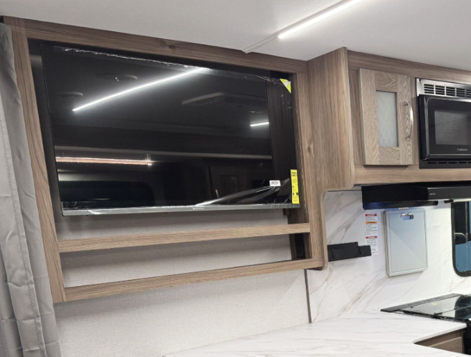 Modern RV interior showcasing a mounted flat-screen TV within a wooden frame, situated above a countertop with a marble-like surface. Adjacent cabinetry includes a built-in microwave and frosted glass storage doors, complemented by under-cabinet lighting for a sleek and functional design.