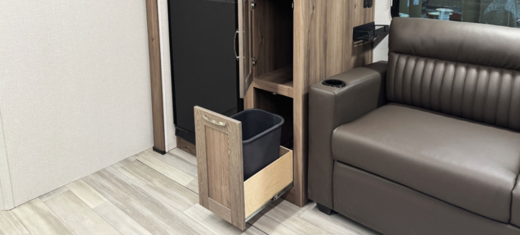 RV interior featuring a brown leather loveseat with a cup holder, next to a wooden cabinet with a pull-out drawer containing a black trash bin. The design showcases light-colored flooring and a compact, organized living space with practical storage solutions.