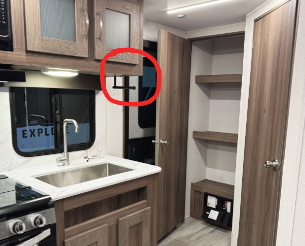 RV kitchen and storage area featuring a sink with a marble-like countertop, a stove, and wooden cabinetry with frosted glass panels. A red circle highlights a wall-mounted hook or hanger near the window. The open shelving and door to a small closet provide additional storage options, with light-colored flooring completing the modern design.