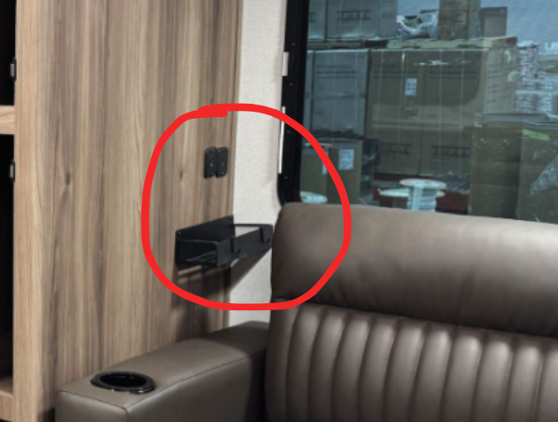 Close-up of an RV living area showing a small wall-mounted black shelf, circled in red, next to electrical outlets. The shelf is located above a brown leather loveseat with a visible cup holder, positioned near a large window with a view of stacked boxes outside.
