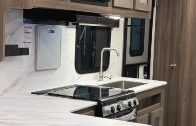 Close-up of an RV kitchen featuring a marble-like countertop, a stainless steel sink with a modern faucet, and a built-in oven with a stovetop. The backsplash and cabinetry have a clean, light wood finish, with frosted glass cabinet doors above. A window over the sink allows natural light into the space, enhancing its sleek and functional design.