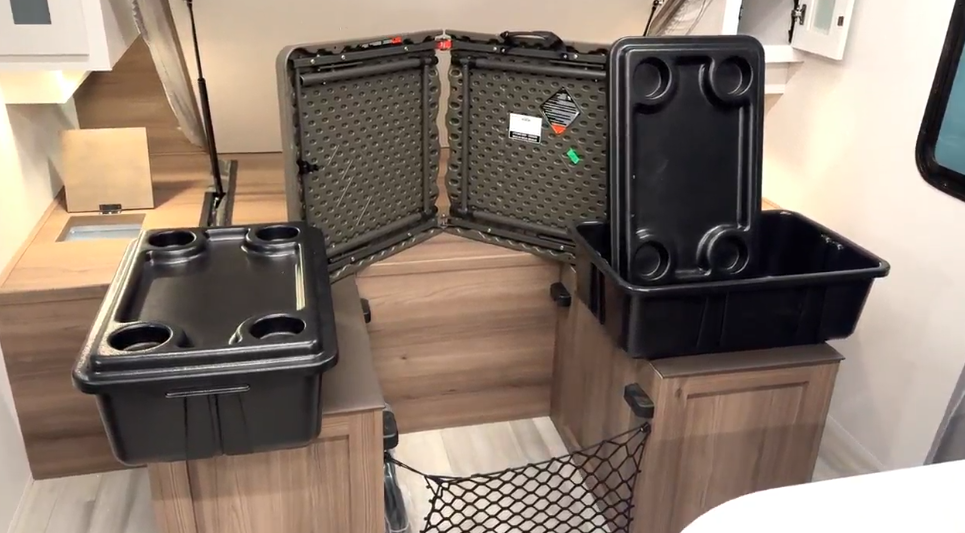 RV interior showcasing storage solutions with two black plastic bins featuring cup holders, one stacked and one open, placed on wooden cabinets. An open hard-shell storage case is propped in the background, and a mesh storage net is visible below. The space highlights practicality and organization within the compact living area.