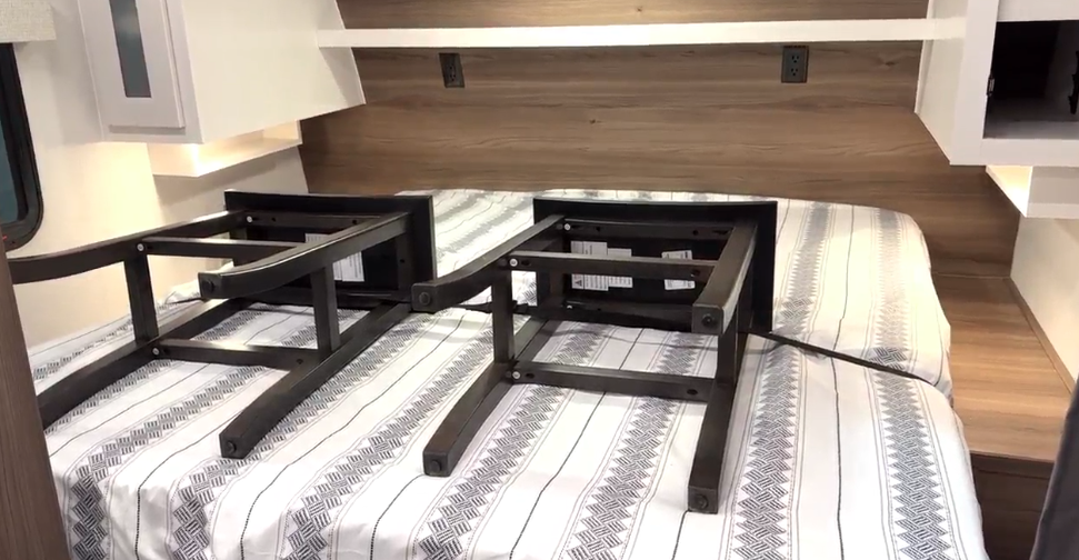 RV bedroom interior featuring a neatly made bed with a striped geometric-patterned comforter. Two black metal table frames are positioned on the bed, surrounded by wooden paneling, white overhead cabinets, and built-in reading lights for a modern and functional design.