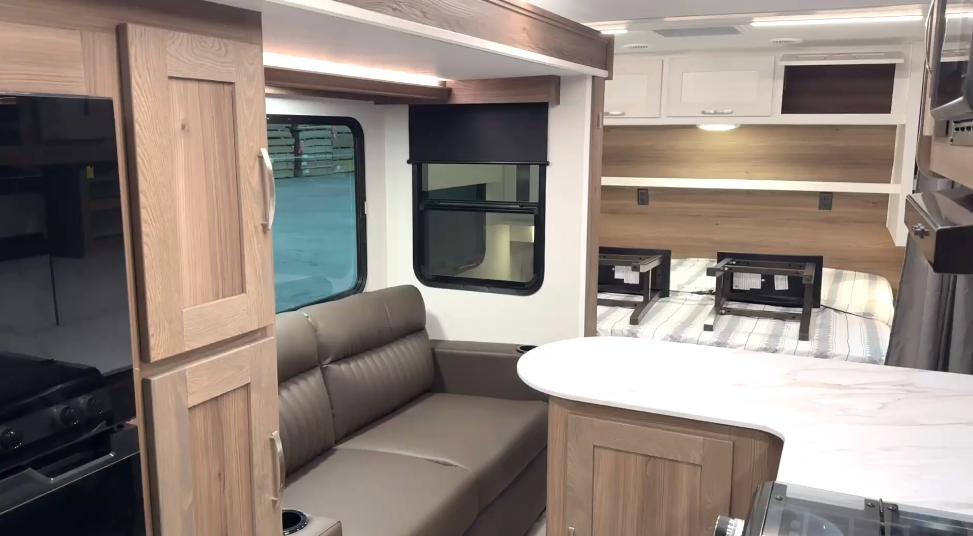 RV interior showcasing an open floor plan with a brown leather loveseat, a kitchen area featuring a marble-like countertop, and a sleeping area with a bed and overhead white cabinets. The bed includes two black storage frames placed on top. Large windows with shades and wooden cabinetry enhance the modern and functional design.