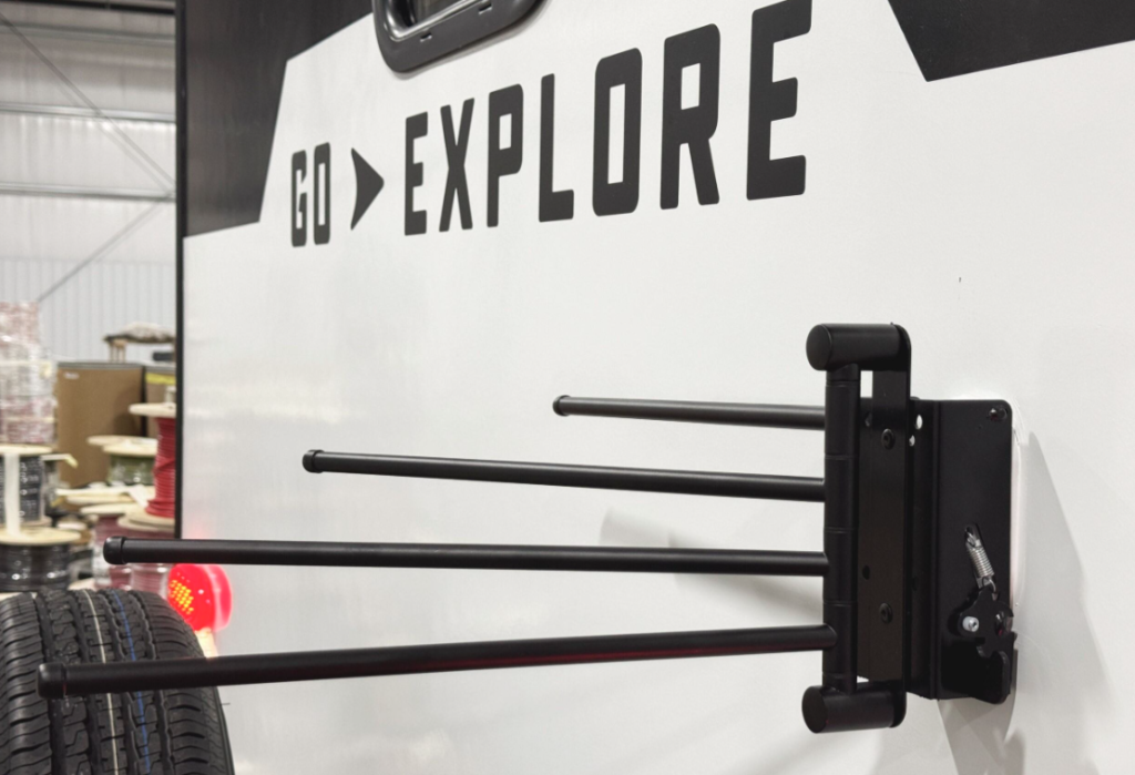 Close-up of the exterior of a travel trailer with the words 'GO EXPLORE' printed on the side. Mounted to the trailer is a black retractable arm system designed for holding items such as towels or gear. A spare tire is partially visible in the foreground, adding to the practical design.