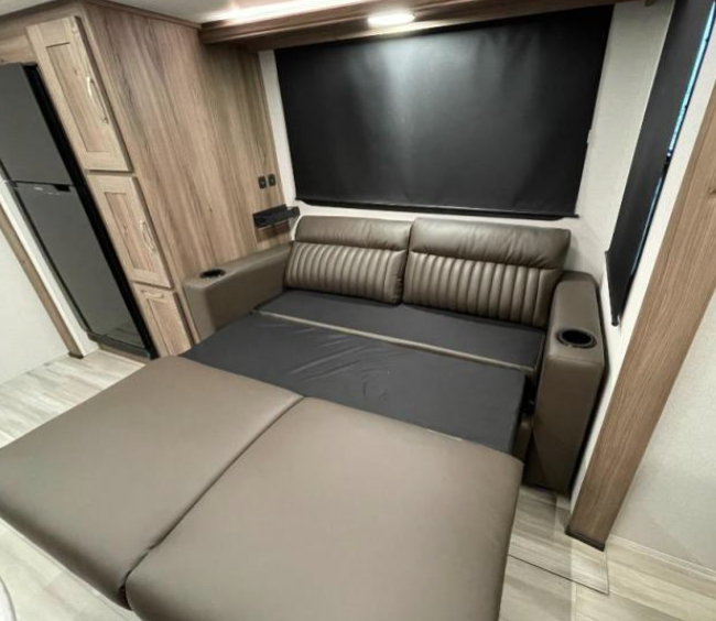 RV interior with a brown leather loveseat unfolded into a sleeper sofa. The space includes wooden cabinetry, a black-out window shade, and a modern, compact layout with light-colored flooring. The sofa bed design adds functionality to the cozy living area.